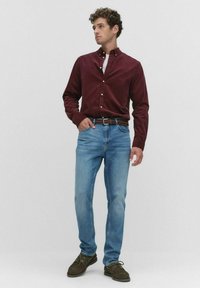 Man in een bordeauxrood overhemd met knoopjes, lichtblauwe jeans met een bruine geweven riem en donkerolijfgroene schoenen, staande tegen een effen achtergrond.