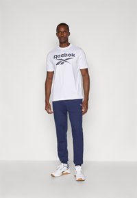 Biela bavlnená tričko s modrým logom Reebok, spárované s námorníckymi teplákmi. Biele športové topánky s gumenou podrážkou dopĺňajú outfit.