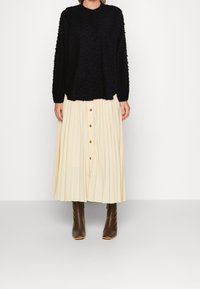 Svart texturerad långärmad topp med knappar, kombinerad med en veckad krämfärgad kjol med framknappar och bruna klackstövlar.