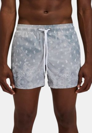Uomo che indossa pantaloncini da bagno grigio chiaro con motivi paisley bianchi e piccoli motivi floreali e vita con coulisse bianca.