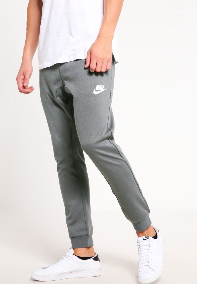 Man som bär grå Nike sweatpants, vita Nike sneakers och en vit t-shirt, stående mot en enkel ljus bakgrund.