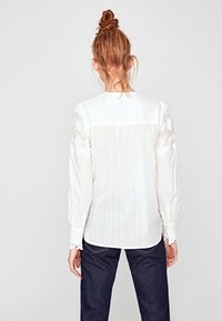 Witte lange mouwen blouse van textuurstof, met kantaccenten op de schouders en gatheren bij de manchetten. Beschikt over plooien aan de achterkant voor een relaxte pasvorm.