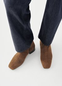 Bottines en daim marron à bout carré, associées à un jean denim bleu foncé, présentant une texture lisse et un talon bloc bas.