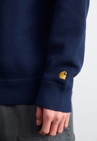 Navy blauwe gebreide trui met geribbelde manchetten en een klein geel logo op de mouw. Het materiaal lijkt dik en is gemaakt voor warmte.