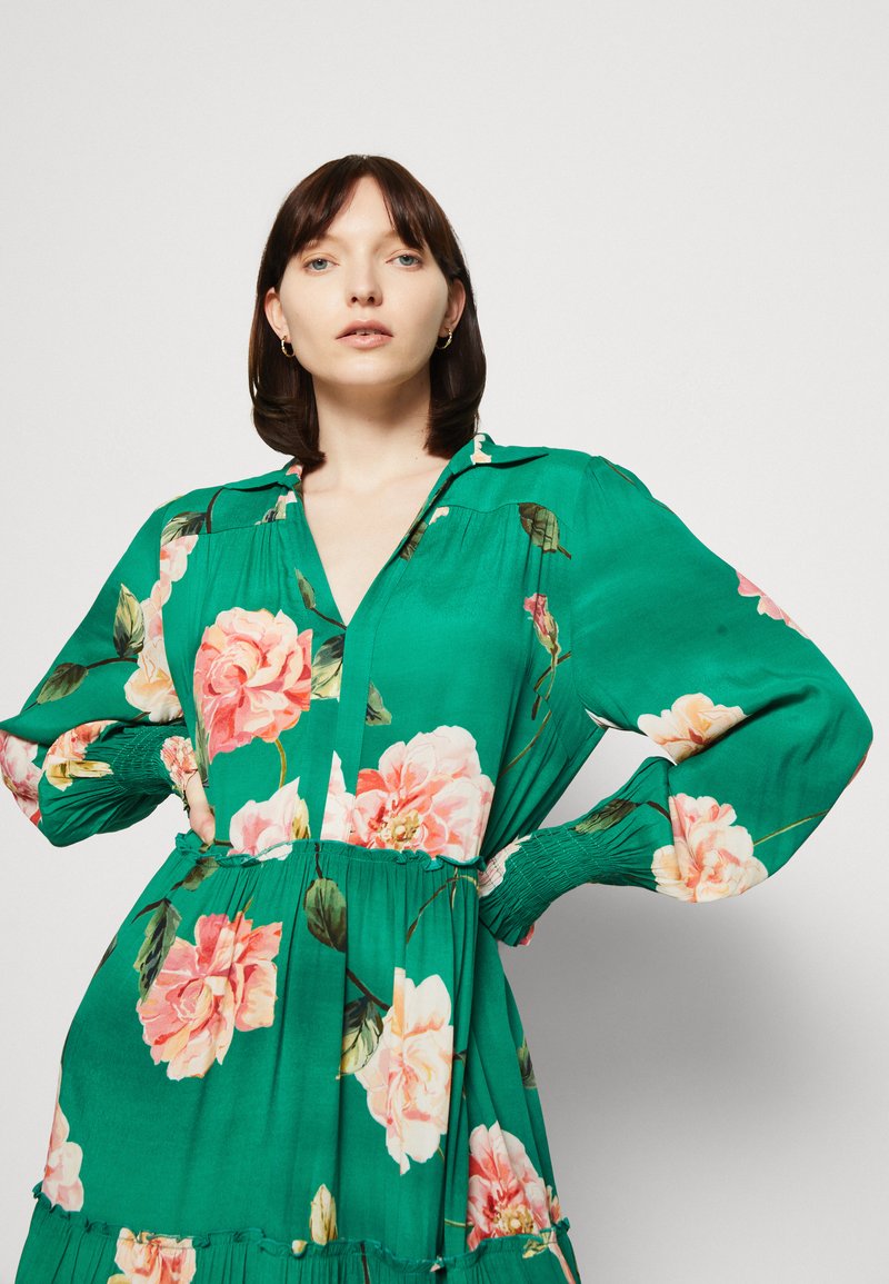 Grünes Blumenkleid mit großen rosa Rosen, langen puffärmeligen Ärmel und V-Ausschnitt. Mit einem gerafften Taillenbereich und einem gestuften Saumdesign.