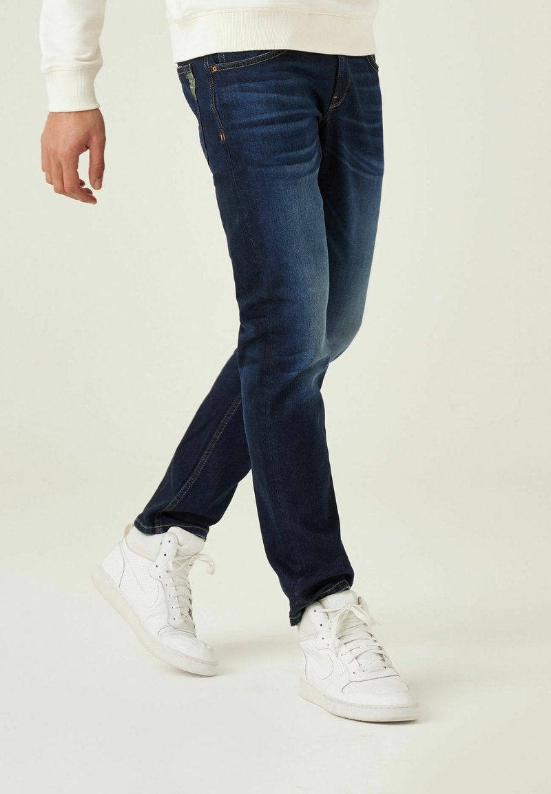 Man i mörkblå jeans och vita högskor, går med ett ben något korsat framför det andra.