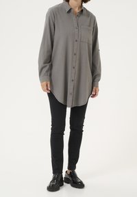 Camisa de botón gris de manga larga hecha de tejido suave, con un bolsillo en el pecho, dobladillo curvado y corte relajado, combinada con pantalones negros ajustados y zapatos.