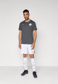 T-shirt grigia Nike con logo, abbinata a pantaloni corti bianchi con branding laterale. Calzature da calcio nere e calze bianche completano l'outfit sportivo.