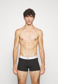 Boxers ajustados negros con una banda elástica blanca que presenta la marca "BOSS" en azul. El modelo tiene una complexión muscular y cabello rizado.