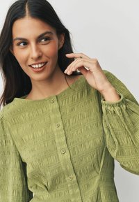 Oliivinvihreä, teksturoitu pusero, jossa leveä pääntie, röyhelöidyt hihat ja napillinen etuosa. Siinä on hienovaraiset pystysuorat raidat ja joustavat yksityiskohdat.