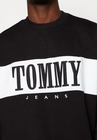 Sudadera en blanco y negro con cuello redondo, que presenta un atrevido logo de "TOMMY" y "JEANS" en texto más pequeño debajo. Textura de tela suave.