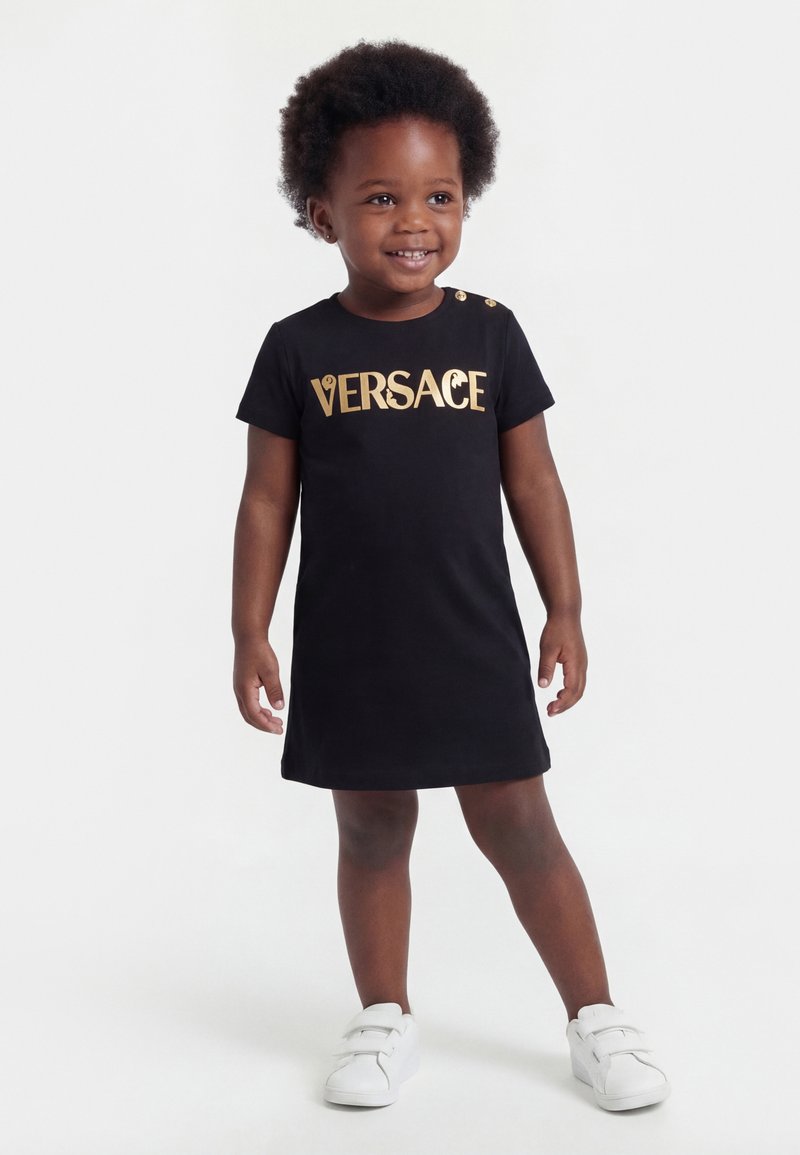 Petite fille souriante portant une robe noire Versace avec des lettres dorées et des baskets blanches, posant devant un fond blanc uni.