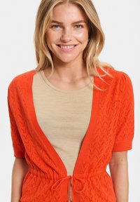 Oranje gebreide cardigan met een v-hals, korte pofmouwen en een koord in de taille, gelaagd over een beige top met korte mouwen.