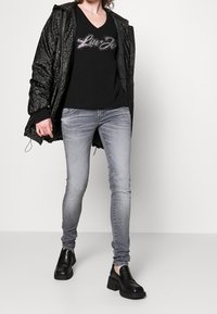 Black puffer jacket with a textured pattern, black v-neck t-shirt with rhinestone detail, light grey skinny jeans, and chunky black shoes.