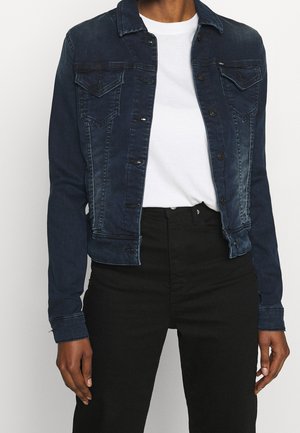 Person iført en mørkeblå denimjakke over en hvid t-shirt og højtaljede sorte bukser, stående mod en ensfarvet baggrund.