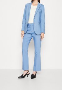 Světlomodrý oblek na míru skládající se z úzkého saka a rovného střihu kalhot, doplněný krémovým topem a černými lodičkami na podpatku.