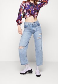 Donna che indossa un top a maniche corte viola a fantasia, jeans strappati azzurri chiari e mocassini platform lavanda con calzini bianchi, in posa con la mano sulla testa.