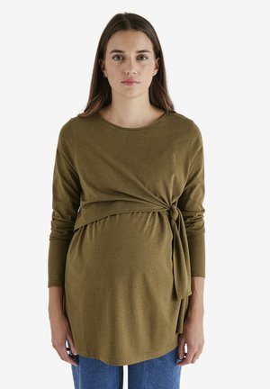Young pregnant woman with long brown hair wearing an olive green long-sleeve top with side tie and blue jeans, standing against a white background.