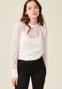 Weißes Spitzenoberteil mit langen Ärmeln und hohem Hals, das ein geometrisches Muster und eine figurbetonte Silhouette aufweist, kombiniert mit schwarzen Jeans.