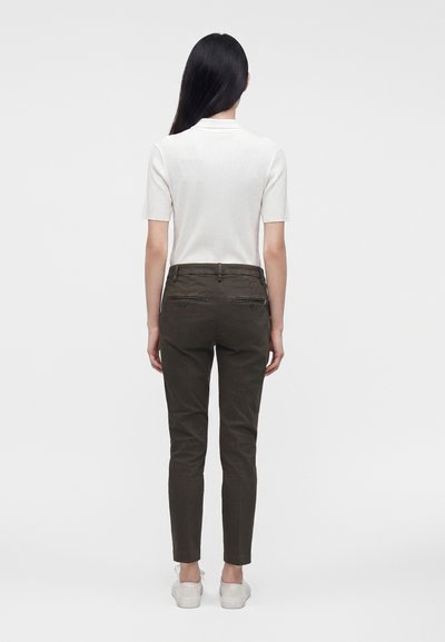 Femme aux longs cheveux noirs portant un haut blanc à manches courtes, un pantalon sombre et ajusté, ainsi que des baskets blanches, debout de dos sur un fond uni.
