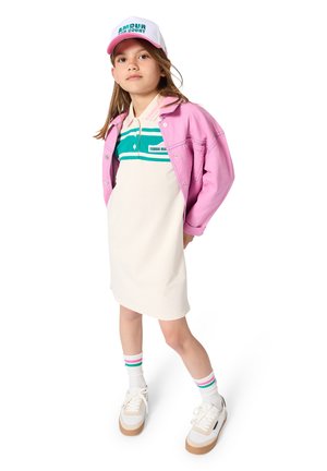 Jeune fille portant une casquette blanche, une veste rose, une robe de tennis crème avec des rayures bleu sarcelle, des chaussettes blanches avec des rayures roses et bleu sarcelle, et des baskets blanches.