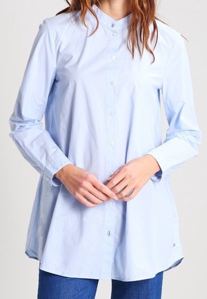 Woman wearing a light blue, long-sleeve button-up shirt with a mandarin collar standing against a plain background.