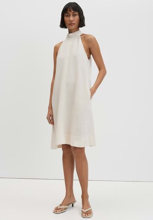 Frau trägt ärmelloses, knielanges, weißes Kleid mit hohem Kragen und Seitentaschen sowie beige offene Kitten-Heel-Sandalen, steht vor einfarbigem Hintergrund.