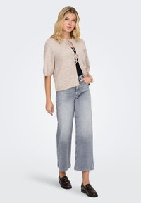 Bež pleten pulover s kratkimi rokavi, zapenjanje na zavezan trak, nošen preko črne majice in kombiniran s širokimi svetlo modrimi kavbojkami ter temnimi loafers čevlji.