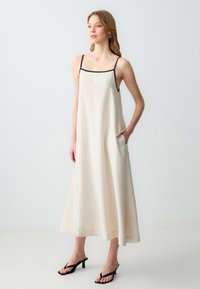 Abito midi senza maniche di colore crema con rifiniture nere sul collo, tasche laterali e una vestibilità comoda. Indossato con sandali neri con cinturini. Tessuto liscio.