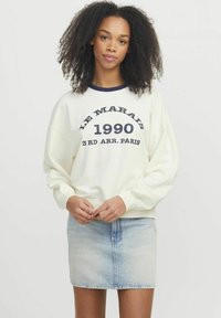 Felpa crema con colletto a coste blu navy e testo stampato. Abbinata a una mini gonna di jeans chiari, che mostra un look casual e stratificato.