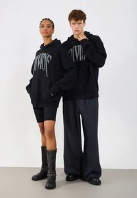 Sudaderas negras con el gráfico "DIVINE" en plateado, combinadas con shorts de ciclista negros y pantalones de pierna ancha. El modelo de la izquierda lleva botas hasta la rodilla.