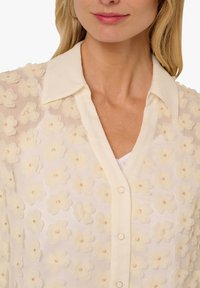 Sheer light beige blouse featuring textured floral appliqués with button closures, a pointed collar, and a smooth inner layer.