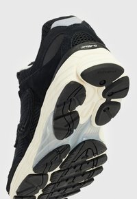 Zapatilla deportiva negra con parte superior de ante y malla, suela texturizada con una entresuela crema y patrones de tracción distintivos en la suela exterior.