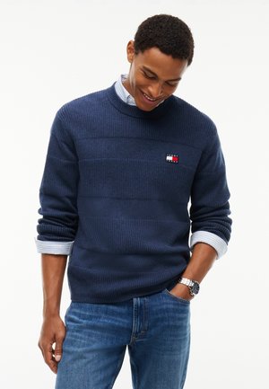 Ung mand iført en mørkeblå sweater over en lyseblå skjorte med krave, blå jeans og et sølvfarvet armbåndsur, smilende og kiggende ned.