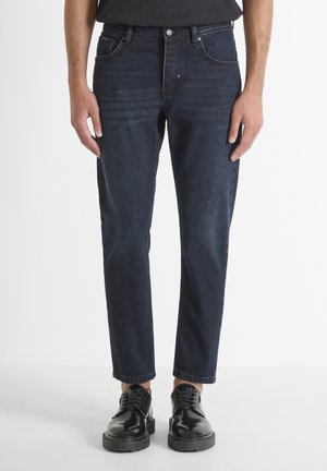 Jeans de mezclilla azul oscuro con corte recto, cinco bolsillos y un ligero desgaste. Combinados con zapatos de cuero negros con cordones para un contraste.