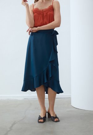 Kvinde iført en ærmeløs rust-rød blondeprydet top, navy blå omslags-midi-nederdel med flæser og sorte hælede sandaler med åben tå, stående indendørs.