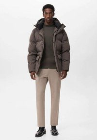 Chaqueta acolchada marrón oscuro con un cuello alto, llevada sobre un suéter verde, combinada con pantalones beige y zapatos negros. Diseño texturizado y cálido.