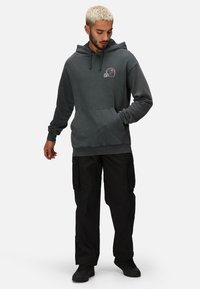 Felpa con cappuccio grigia antracite con tasca frontale, dotata di una piccola grafica di un casco sportivo, abbinata a pantaloni cargo neri e scarpe.