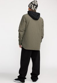 Veste à manches longues vert olive avec une capuche noire, texture côtelée, boutons aux poignets, associée à un pantalon noir large et des baskets noires.