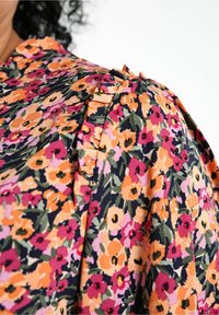 Donna che indossa una blusa floreale con fiori arancioni, rosa, viola e foglie verdi, con dettagli plissettati sulle spalle.