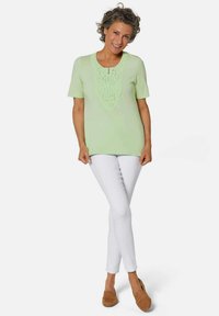 Top verde chiaro a maniche corte con dettagli in pizzo testurizzato nella parte anteriore, abbinato a pantaloni bianchi aderenti e mocassini marroni. Design semplice.