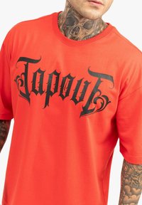 Sarkans kokvilnas T-krekls ar lielu melnu "Tapout" logotipu un dekoratīviem ziedojumiem. Īsas piedurknes un apkakle ar apaļu izgriezumu.