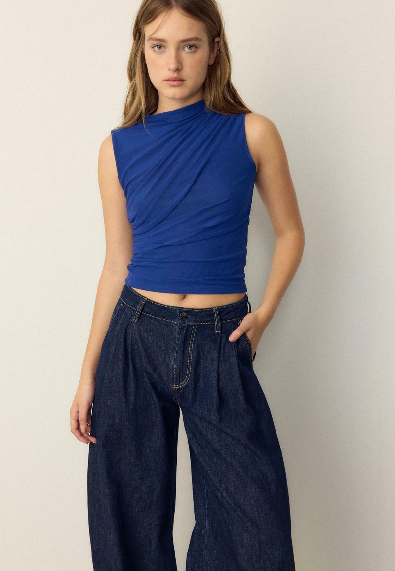 Junge Frau trägt ein ärmelloses, königsblaues Oberteil mit Raffungen und locker sitzende, dunkle, weit geschnittene Jeans, steht vor einem einfachen, hellen Hintergrund.
