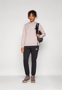 Világos rózsaszín kapucnis pulóver logóval, fekete melegítőnadrág és barna tornacipő. A modell egy fekete hátizsákot tart, göndör haja pedig körbeöleli az arcát.