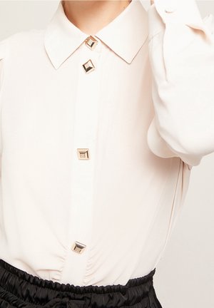Jasnoróżowa bluzka z klasycznym kołnierzem, ozdobiona dużymi, kwadratowymi złotymi guzikami i delikatnym marszczeniem w talii. Miękka tekstura materiału.