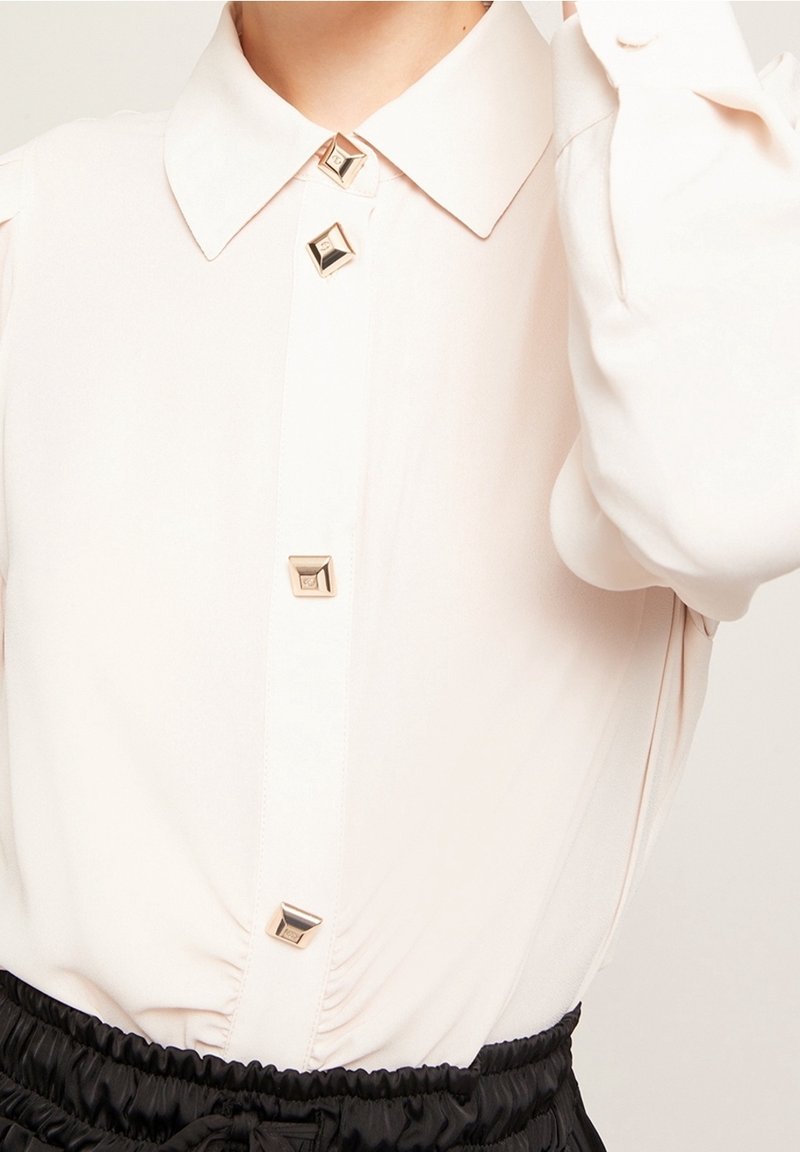 Blouse rose clair avec col classique, ornée de grands boutons carrés dorés et d'un léger froncé à la taille. Texture de tissu douce.