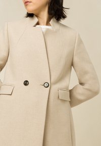 Beige jas van textuurstof, met een hoge kraag, dubbele knopen sluiting en twee voorzakken.