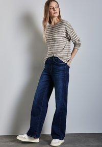 Licht beige en donkergrijze gestreepte blouse met lange mouwen, gecombineerd met donkerblauwe wijde jeans en witte sneakers, tegen een effen achtergrond.