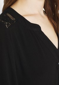 Blusa nera con scollatura a V, dettagli in pizzo sulle spalle e tessuto morbido e fluente con un delicato drappeggio.