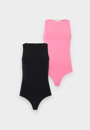 Kvinne med bølget hår iført svart ermeløs topp, vide blå jeans og svarte joggesko, stående foran en ensfarget hvit bakgrunn.